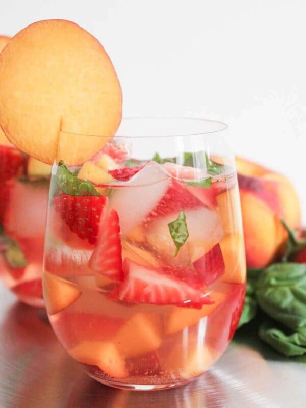 Two glasses of sparkling rose sangria garnished with thin peach slices in front of a display of summer fruits.