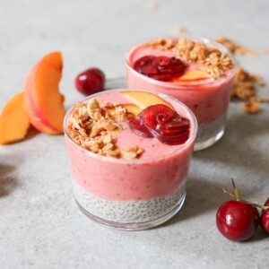 Two glasses of peach-cherry smoothie and coconut chia seed pudding topped with granola, cherry, and peach slice.