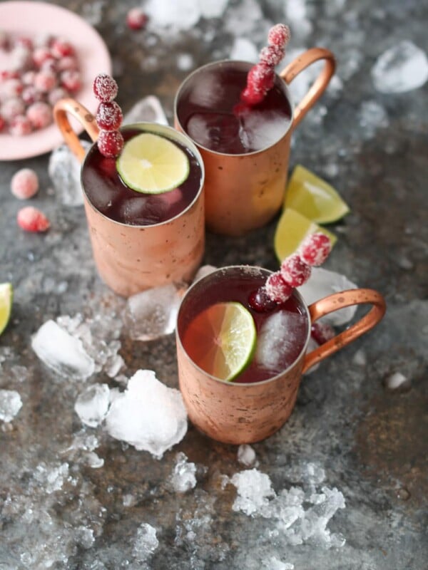 Overhead shot of three cranberry moscow mules in copper mugs with sugared cranberry swizzle sticks and bowl of sugared cranberries.