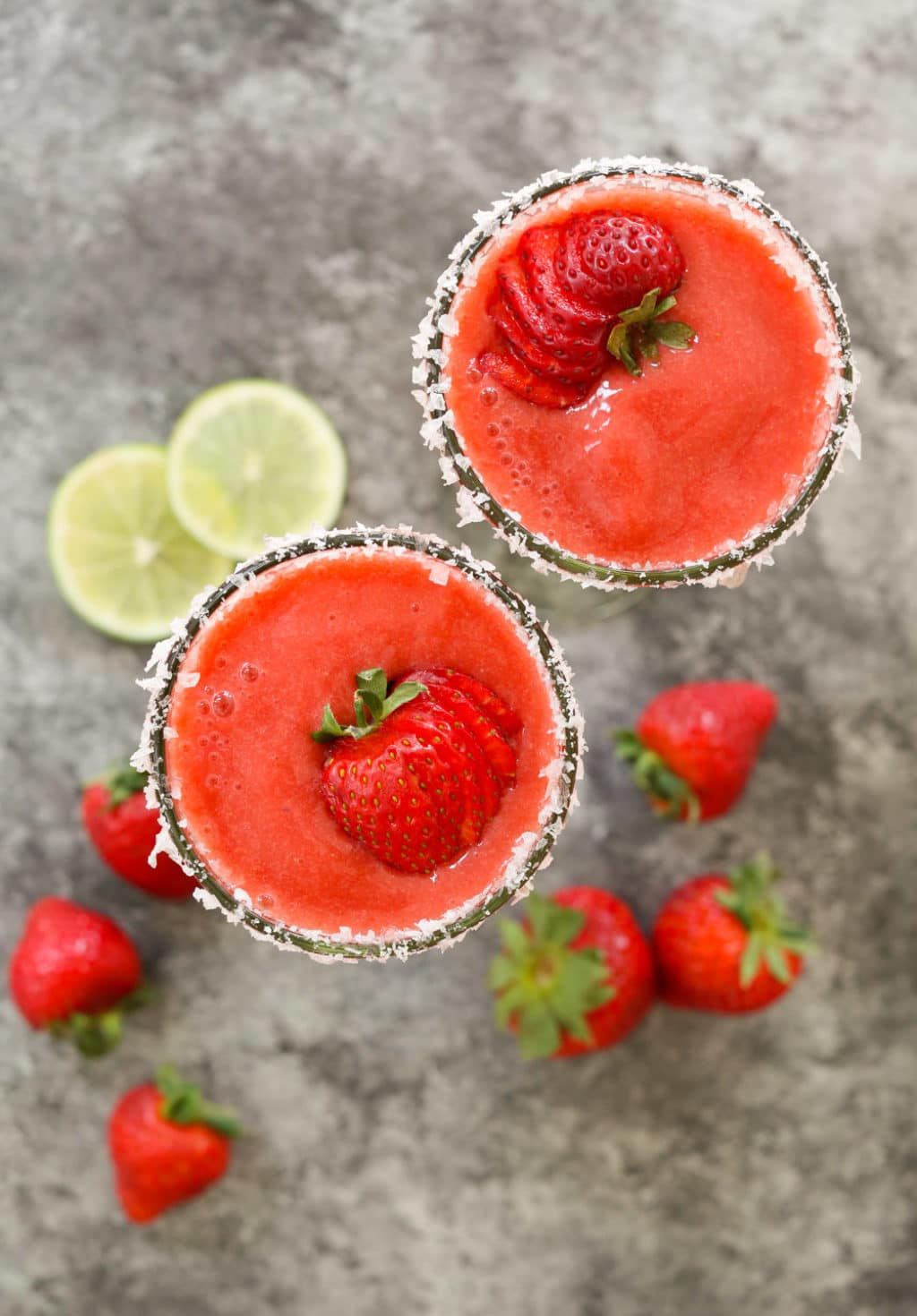 Overhead shot of two frozen margaritas with salted rims and sliced strawberry garnish.