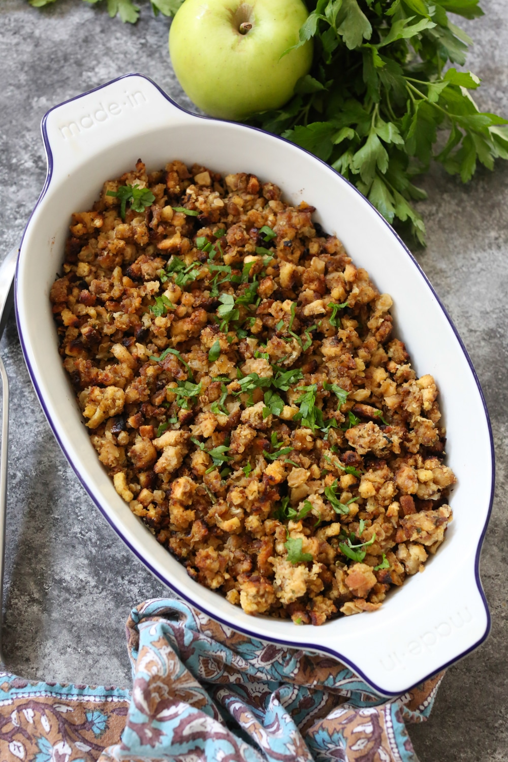 Side view of Stovetop Stuffing with Sausage, Apple and Onion in an oval baking dish with a serving spoon to the left and some parsley and apple peeking into the frame.